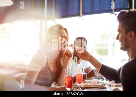 Two young couples having fun, feeding each other with slice of pizza, group of multiracial friends sharing pizza together. Food, Friendship and Togeth Stock Photo