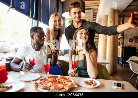 Multiethnic group of young cheerful students having fun at cafe on the party with tasty fast food, drinks, taking selfie on smartphone Stock Photo