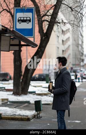 a young man is standing at the bus stop and waiting for the bus. He holds a backpack and drinks coffee from a reusable Cup. The guy looks forward with Stock Photo