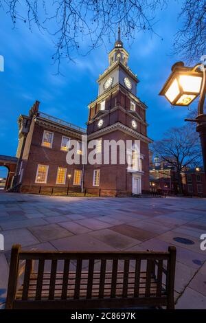 Philadelphia, PA--Feb 4, 2020; lights in the windows and clock tower of Independence Hall illuminate the grounds with wooden bench and street lamp in Stock Photo