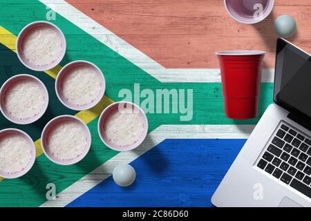 South Africa flag concept with plastic beer pong cups and laptop on national wooden table, top view. Beer Pong game. Stock Photo