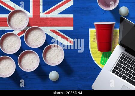 Turks And Caicos Islands flag concept with plastic beer pong cups and laptop on national wooden table, top view. Beer Pong game. Stock Photo