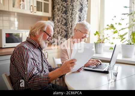 Content senior bearded greyheaded husband and his wife smiling checking utility bills or insurance at computer with easy access, sitting at kitchen ta Stock Photo