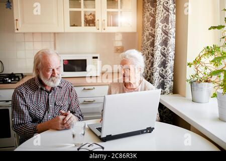 Handsome cheerful senior couple sitting at the kitchen table and using digital tablet for paying utility bills. Senior lifestyle, mature people and te Stock Photo