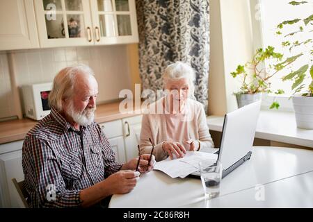 Content senior bearded greyheaded husband and his wife smiling checking utility bills or insurance at computer with easy access, sitting at kitchen ta Stock Photo