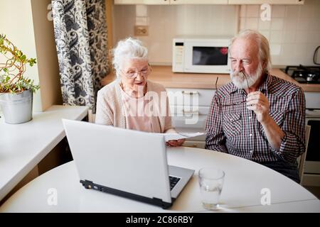 Senior couple at home kitchen using laptop, writing email to their foreign far-away friends, one of the pensioners holding document, another typing on Stock Photo