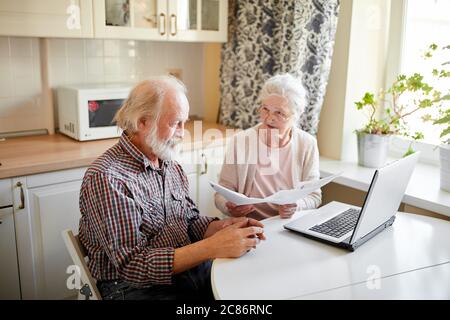 Mature couple sitting at kitchen table with laptop looking through financial papers, having little jam with the pension contributions. Stock Photo