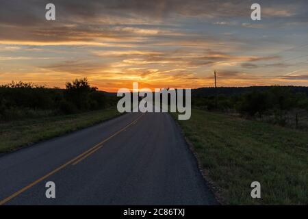 Rural two lane country road curving slightly as it turns to head toward a beautiful sunset with fiery orange and yellow clouds on the distant horizon. Stock Photo