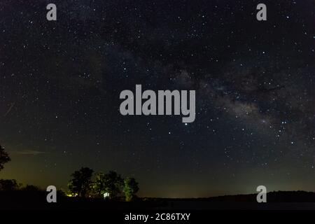 Trees in a campsite on a lake shore lit up by lights in the camp site with the Milky Way in the night sky overhead. Stock Photo