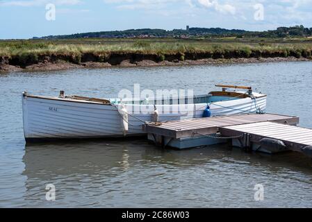 Morston Quay, Norfolk, UK. White wooden clinker built sailing dingy Seal at moorings. The village of Blakeney and the tower of Blakeney Church visible in the distance. Credit: Stephen Bell/Alamy Stock Photo