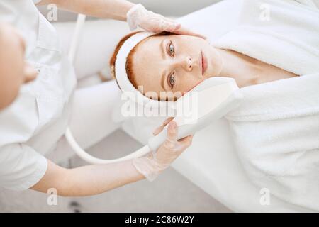 Cosmetology RF lifting equipment. Anti-age and anti-wrinkle apatherapy. Top view of beautiful woman in white bathrobe getting facial treatment and loo Stock Photo