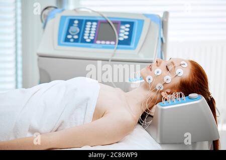Young woman lying with electrodes on her face, receiving electric stimulation on skin. Professional equipment for microcurrent lift face. Anti aging f Stock Photo