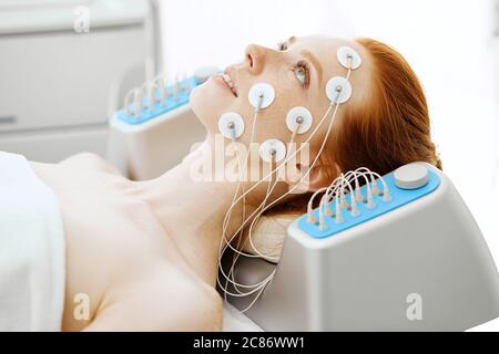 Young woman lying with electrodes on her face in beauty clinic, receiving electric stimulation on skin. Professional equipment for microcurrent lift f Stock Photo