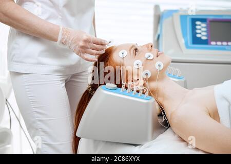 Young woman lying with electrodes on her face, receiving electric stimulation on her skin. Professional equipment for microcurrent lift face. Anti agi Stock Photo