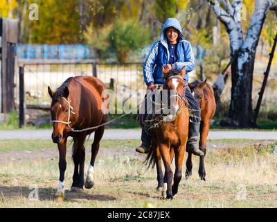 Middle age countryside man in Kyrgyzstan riding a horse wearing a blue jacket with hood. Horseback in rural area near Issyk Kul Kyrgyz Region. Stock Photo