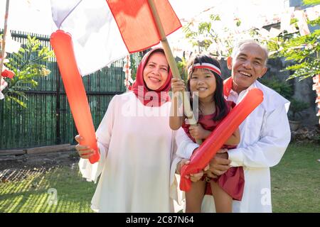 Asian little girl raises the Indonesian flag with a stick while with her grandparents celebrating Independence Day in the yard Stock Photo