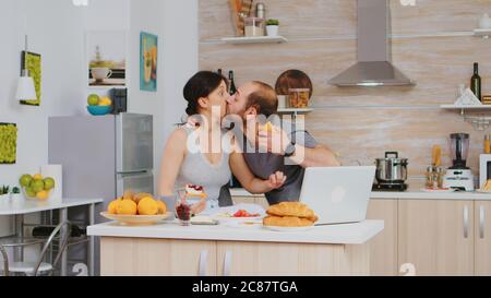 Entrepreneur late at work during breakfast with wife in kitchen. Stressed out man late hurrying nervous running fast to meeting, hurry to work, late for appointment. Kissing wife goodbuy and running to office Stock Photo