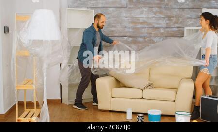 Couple wrapping sofa in plastic foil for protection while they are renovate living room. Home renovation, construction, painting work. Stock Photo