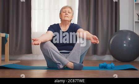 Senior woman meditating sitting on yoga mat in living room. Active healthy lifestyle sporty old person training workout home wellness and indoor exercising Stock Photo