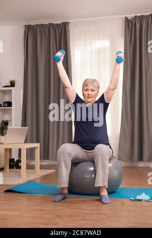 Elderly woman exercising with dumbbells to lose weight. Old person pensioner online internet exercise training at home sport activity with dumbbell, resistance band, swiss ball at elderly retirement age Stock Photo