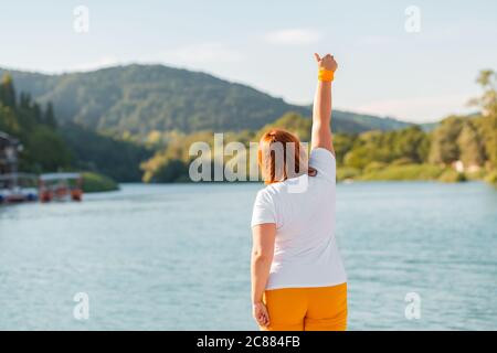 Success. The overweight woman holds up her right hand and gives a thumbs-up. The view from the back. Copy space. Stock Photo
