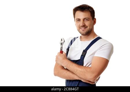 smiling caucasian repairman in blue overalls holds adjustable wrench. worker with arms folded isolated on white studio background. copy space Stock Photo