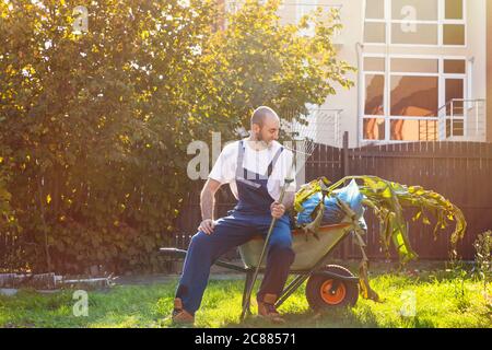 The gardener sits on the cart with leaves. The sun is shining brightly. Smile and good mood. Gardening and cleaning. Stock Photo