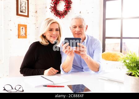 Shot of mature couple sitting at desk at home and making a video call while using mobile phone at home. Stock Photo