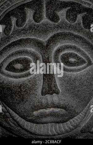 Crudely carved face on bowl of baptismal font in Anglican Church of St Thomas at Overmonmow, in Monmouth, Monmouthshire, Wales, UK. Experts used to agree that this and other carvings on the font dated from the 11th or 12th century, but some modern writers believe the ‘Romanesque’ carvings are a 19th century pastiche.  The Welsh historic environment service describes the supporting stem as 19th century, with a “charming possibly recut Romanesque bowl”. Stock Photo