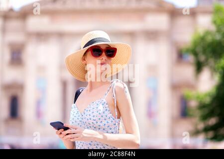 Shot of young woman wearing sunglasses and straw hat and text messaging while walking in the city. Stock Photo