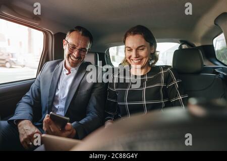 Businesswoman working on laptop with a businessman using mobile phone while traveling by a car. Business people working in backseat of taxi. Stock Photo
