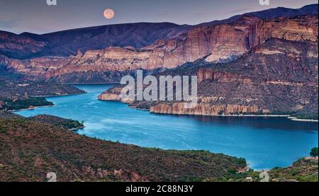 Moon setting down at sunrise over Apache Lake in Superstition Mountains, view from Apache Trail, Arizona, USA Stock Photo