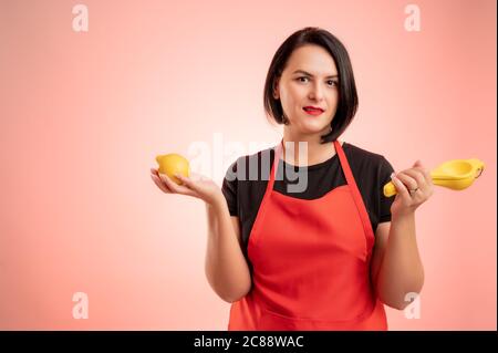 Woman employed at supermarket with red apron and black t-shirt, hold a lemon and a lemon juicer in her hand isolated on red background Stock Photo