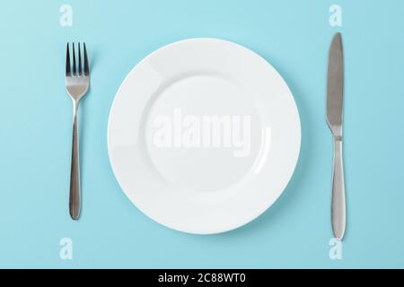 Empty white dinner plate, table knife and fork on a blue background. Set table. Copy space on dish for food design. Top view. Stock Photo