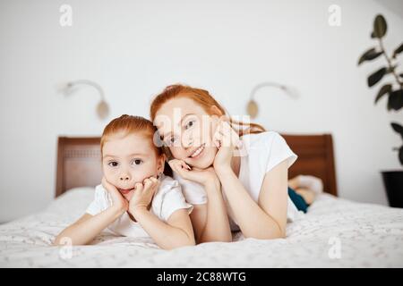 Young red-haired mother in home wear with her little four year old daughter looking at camera while lying on bed in white bedroom. Enjoying free time Stock Photo