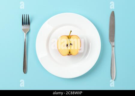 Half of ripe juicy apple on a white dinner plate, table knife and fork on a blue background. Concept of healthy eating, vegetarianism, raw food diet. Stock Photo