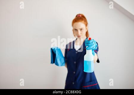 Positive pretty young cleaning lady in blue uniform holds cleaning tools and shows spray bottle. Stock Photo