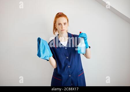 Beautiful red-haired housewife wearing household rubber gloves and blue dressing gown looking at camera posing with blue dusting cloth and wiper. Home Stock Photo