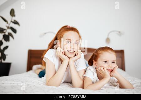 Young red-haired mother in home wear with her little four year old daughter looking at camera while lying on bed in white bedroom. Enjoying free time Stock Photo