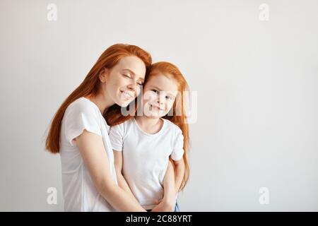 Close-up portrait of smiling mother hugging cute four year old daughter isolated on white background. Two red headed similar females of different gene Stock Photo