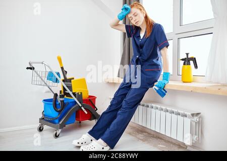 Tired red-haired hotel maid dressed in professional blue uniform in the end of her working day, leaning on window sill wipes out sweat from her forehe Stock Photo
