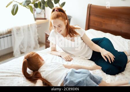 Young red-haired mother in home wear talking with her little four year old daughter while lying on bed in white bedroom. Enjoying free time together w Stock Photo