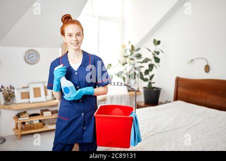 Beautiful red-haired housewife wearing household rubber gloves and blue dressing gown looking at camera posing with red cleaning backet and wiper. Hom Stock Photo