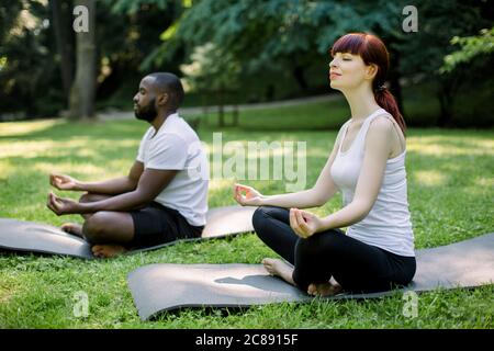 Young multiethnic friends couple doing sport and yoga outdoors, sitting on yoga mats in park in lotus pose. Yoga in park during morning sunrise Stock Photo