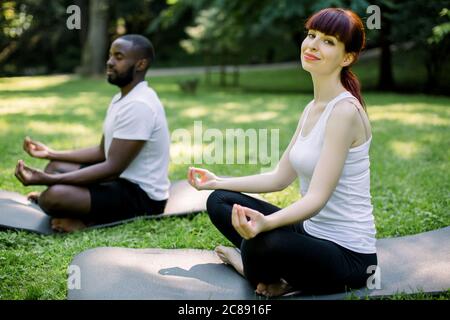 Multiethnic couple, smiling attractive Caucasian woman and concentrated African man, doing yoga in nature, sitting on yoga mats in lotus position Stock Photo