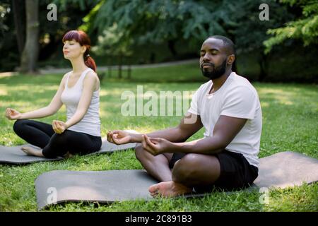Side angle view of good looking young multiethnic couple, African man and Caucasian woman, meditating and doing some yoga together at a park, sitting Stock Photo