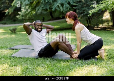 Personal trainer, pretty smiling Caucasian girl, showing abdomen crunches exercise to her male client, joyful African guy. Multiethnical couple doing Stock Photo