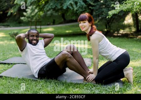 Fitness, sport, training, teamwork and people concept. Handsome muscular African man, doing sit ups in green summer park with his personal trainer Stock Photo
