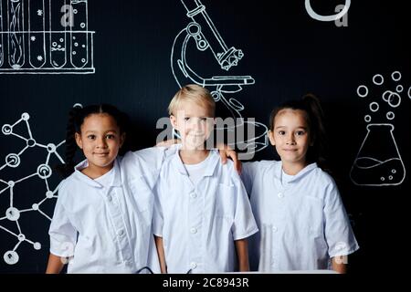 Multiracial group of diverse cute little children in white wear posing against black board with drawings at lab, two mixed race girls hugging a blonde Stock Photo