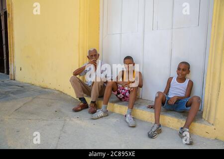 Havana / Cuba - 04.15.2015: Old Cuban man and kids sitting on the street in front of a yellow building; old man smoking a cigar in the old town, Havan Stock Photo